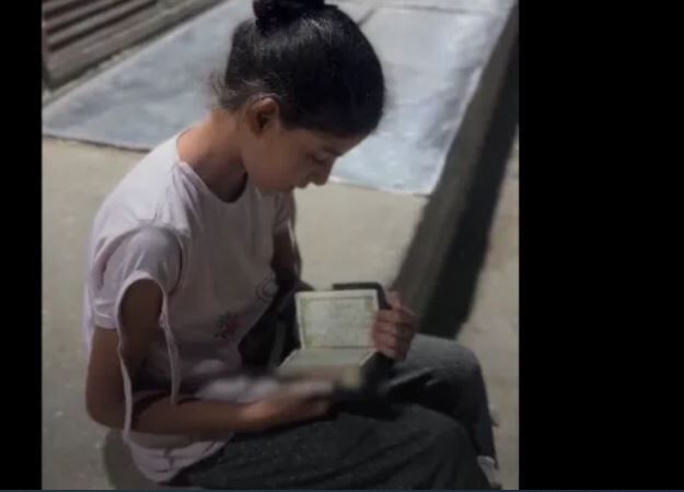 یک دختر فلسطینی در حال تلاوت قرآن مقابل سردخانه‌ای در غزه